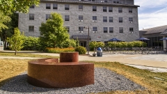 a smoothly polished half-circle bench and cylindrical table, both made of red granite, sit on a patch of gravel in the foreground with a large stone dormitory in the background