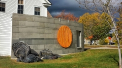 a sprawling sculpture made of discarded tires and colorful insullation leans against a brushed metal wall with dark skies and bright foliage behind