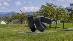 a serpentine aluminum sculpture, painted black, balances atop a bed of small stones surrounded by grass, set against a backdrop of green trees, mountains, and blue skies with hazy clouds