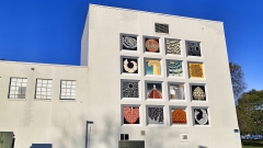 a mural comprised of a grid of 16 squares each with a colorful graphic, on the white wall of a building with blue sky above