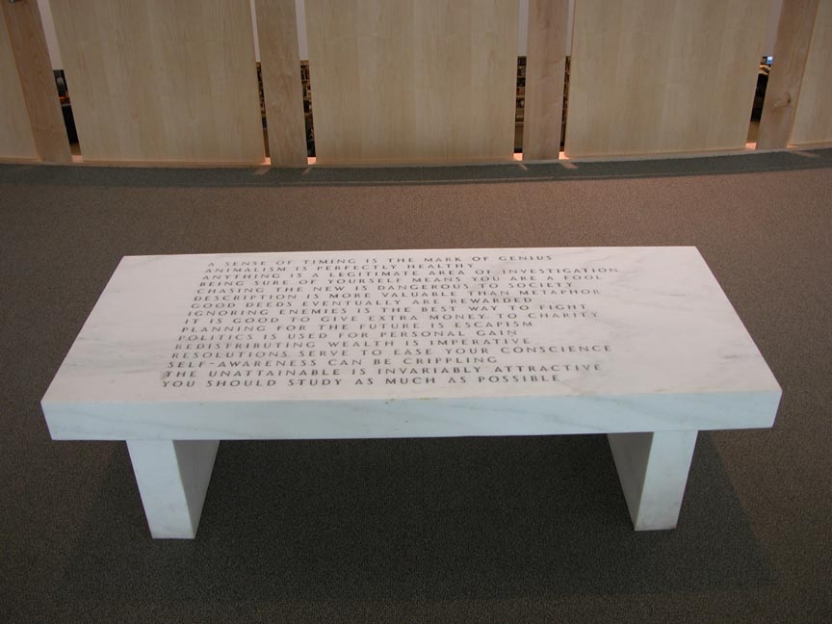 a white marble bench with words carved into it, sits on an expanse of brown low-pile carpet with a tall wooden railing behind