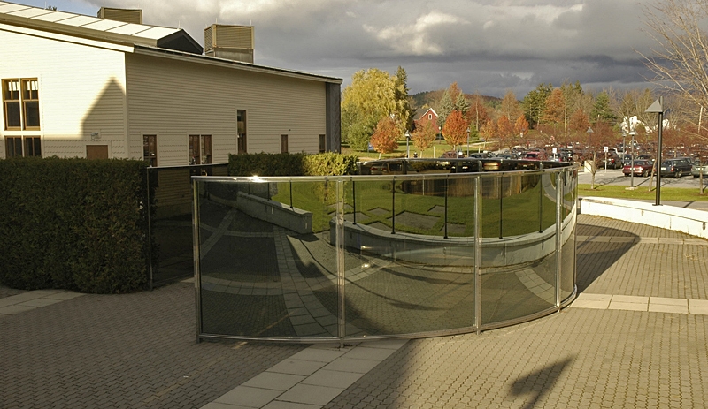 a serpentine sculpture of steel and two-way mirrored glass with arbor vitae elements on an open plaza with fall foliage in the background