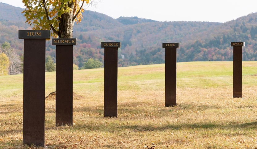 five steel pillars each topped with a dark gray slate placard bearing a word carved and painted gold; words include "HUM," "MURMUR," "GURGLE," "CLICK," and "CRACKLE"