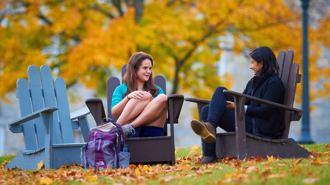 Two students sitting in chairs with the fall foliage in the background