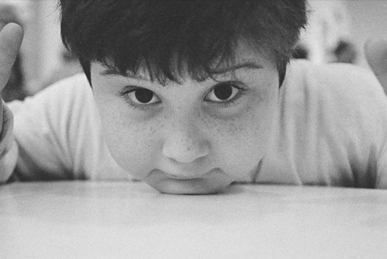 Black and White photo of a child resting their chin on a table looking directly into the camera