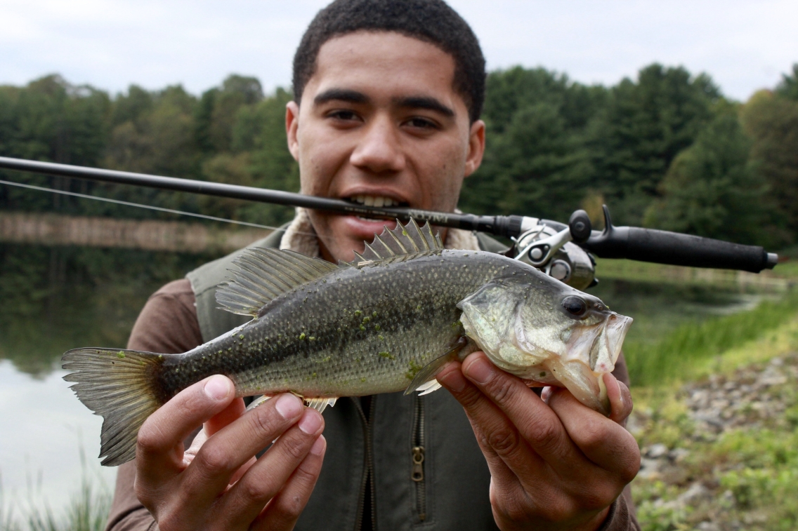 LeeCharles holds up a large green fish while holding his fishing rod in his mouth.