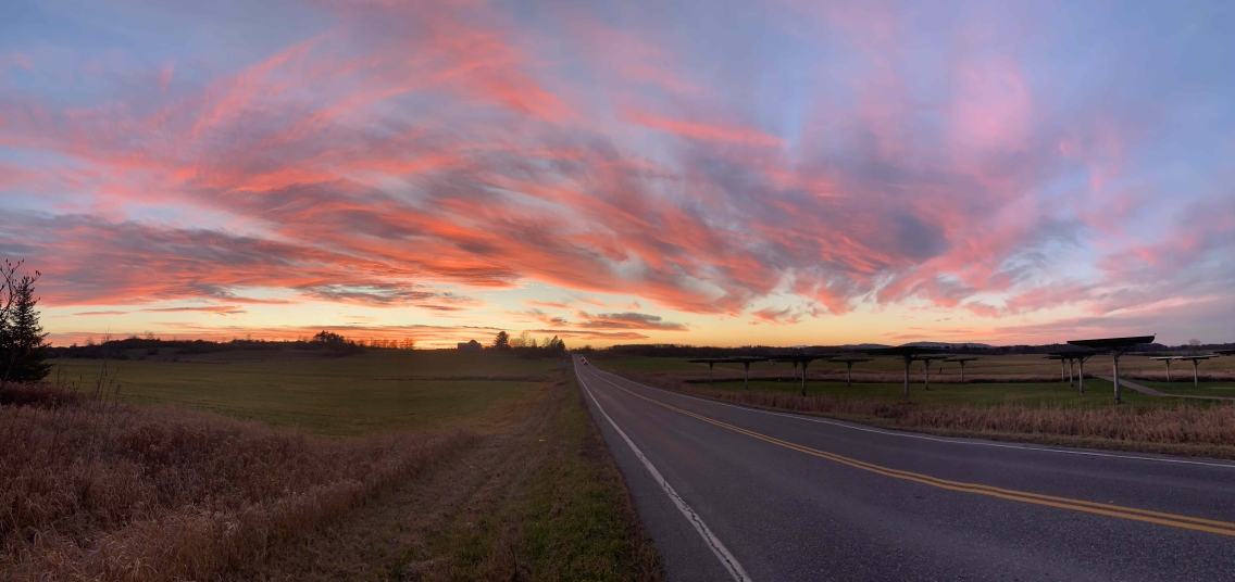 An orange and blue sunset hovers over a long stretch of road and grass.