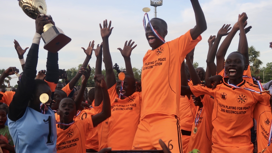 A group of soccer players in bright orange jerseys hoist a trophy and celebrate their victory