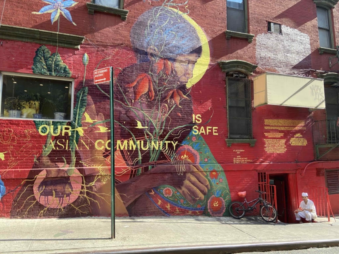 A mural on a red building depicting a woman in a multi-colored outfit with the yellow words "Our Asian Community is Safe" written around her.