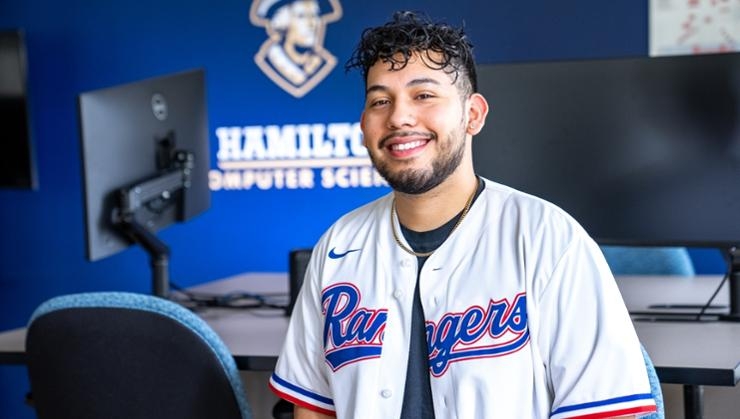Adam Valencia smiles for the camera in a white jersey with text "Rangers" in blue. A computer lab with black monitors and grey desks extends behind him.