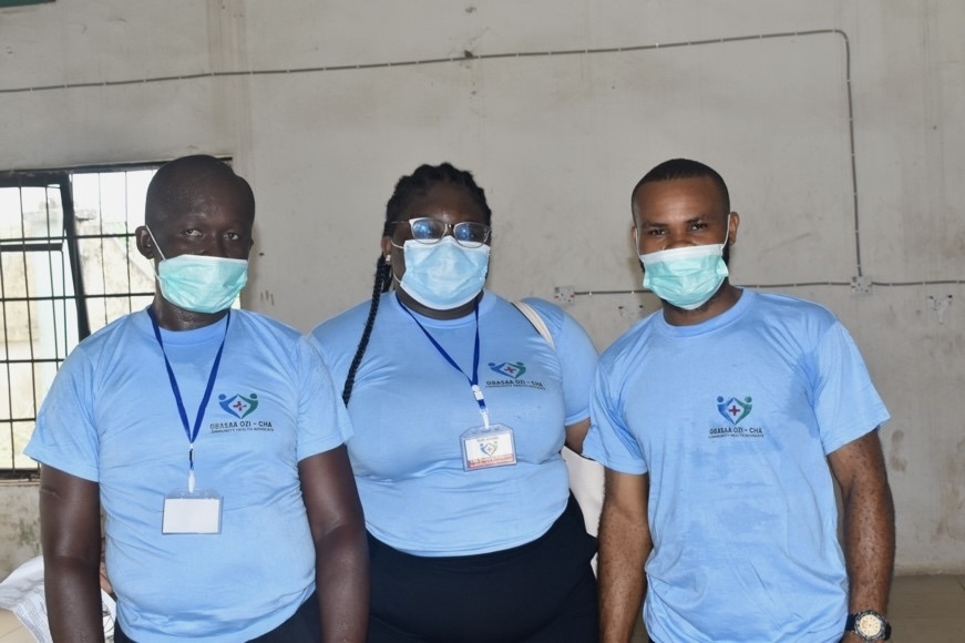 Three people in light blue t-shirts and surgical masks pose for a photo in front of a white wall.