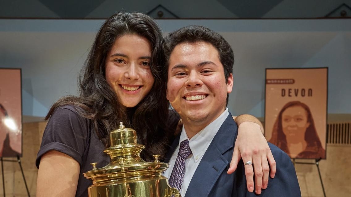 Two students stand with their arms around each other while jointly holding up a gleaming gold trophy on a stage.