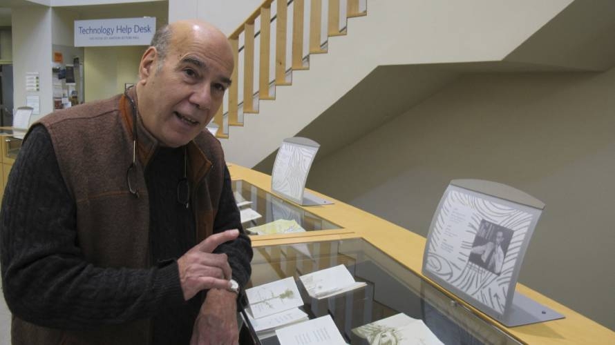 Jay Parini, wearing a brown vest and black shirt, leans on a glass and wood display case filled with Christmas cards.