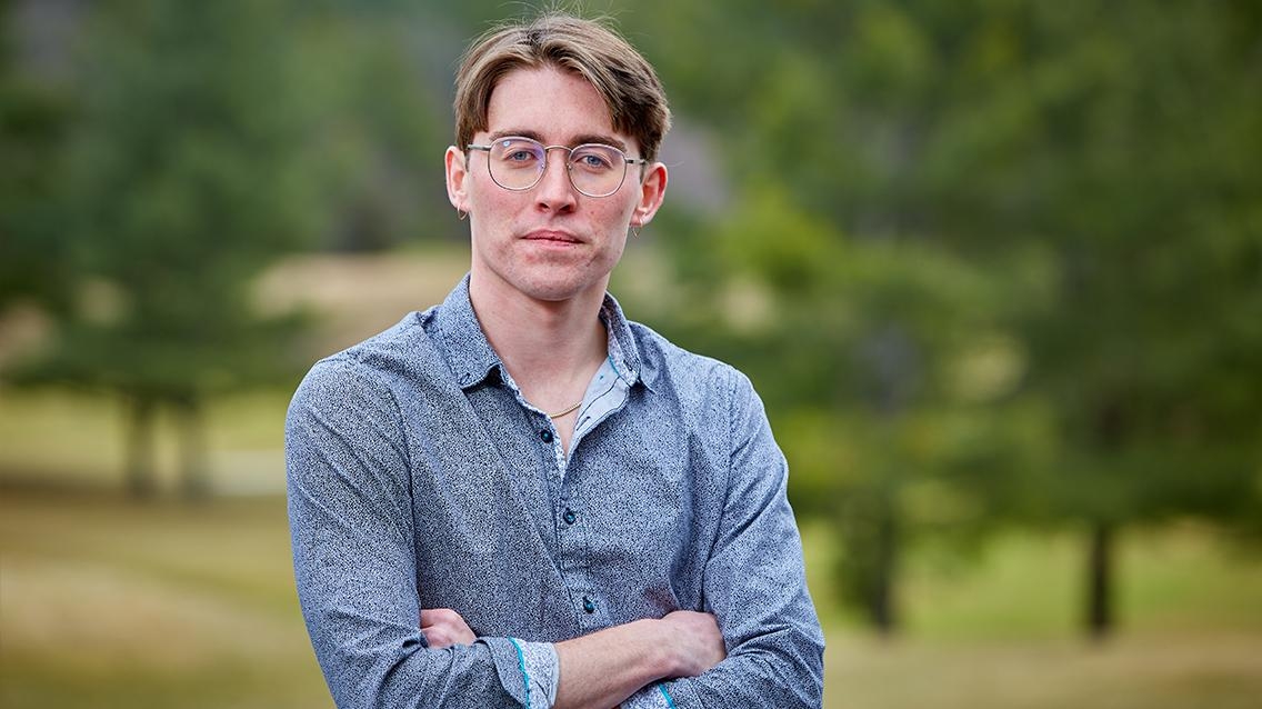 Conor Wertz wears a blue button down and glasses with his arms crossed, posing in front of a stand of green trees.