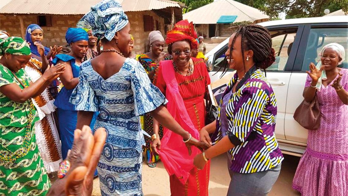 A group of women with dark skin and dressed in colorful dresses and headscarves dance around outside.