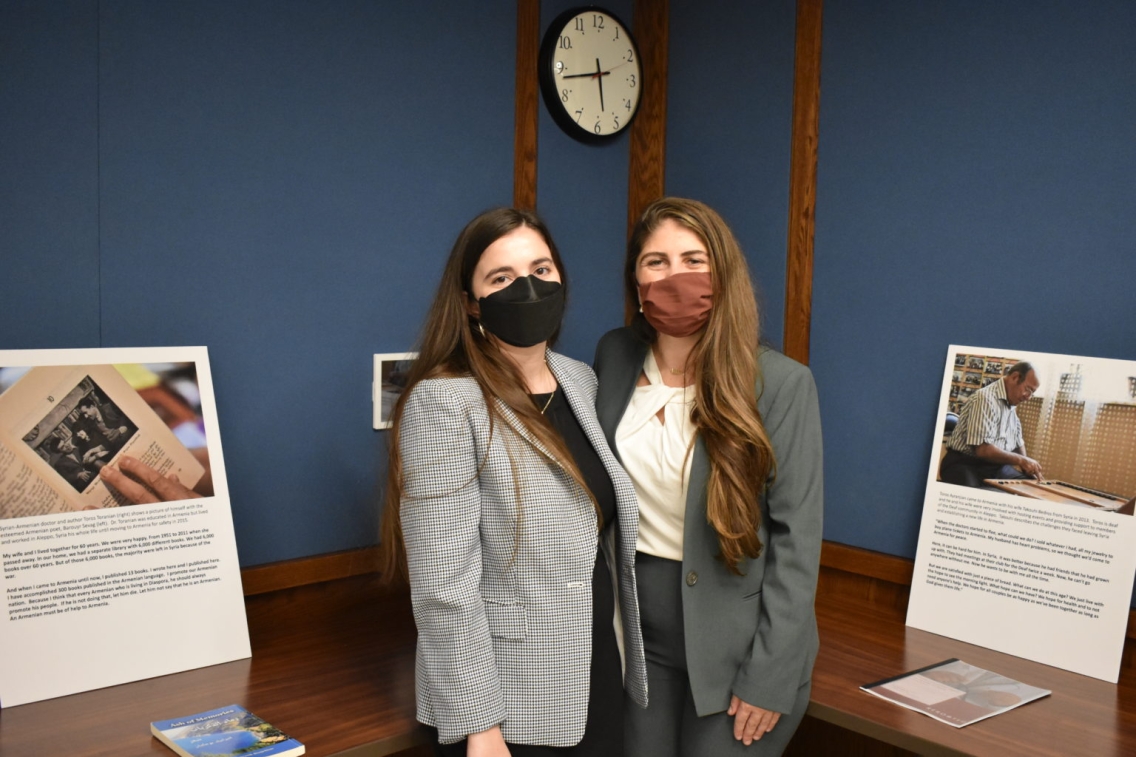 Ani and Anoush pose together, wearing masks and suits, in front of a blue wall and flanked by informational posters.