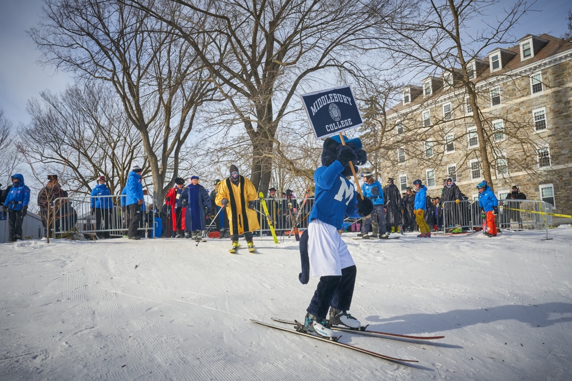 A person in the Middlebury Panther mascot suit skis down a hill while holding a blue and white Middlebury College sign, flanked by graduates in robes ready to follow them down.