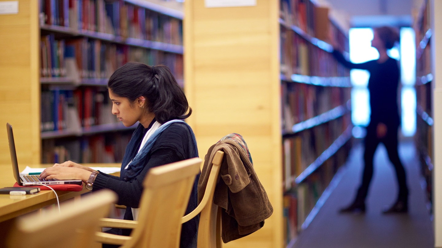 Student sits at a desk in the Davis Family Library working on her computer, while another student browses a bookshelf behind her