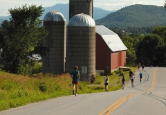 People running in a race on an open road in front of a barn and silo