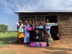 A group of women pose in a group photo holding multi-colored bags they have woven, and stand in front of a wooden cottage.