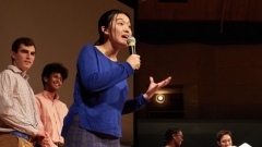 A student in a dark blue shirt and gray pants holds a microphone and delivers a speech on a darkened stage flanked by watching peers.
