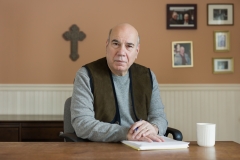 Wearing a gray shirt and brown vest, Jay Parini sits at a light wooden desk with his hands folded on a white notebook next to a white coffee cup.