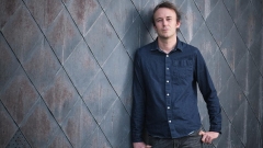 Ross McIntosh, wearing a blue button-down and black pants, leans against a gray, tiled, wall and poses for a portrait.