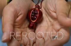 Two hands holds a red pendant shaped like a pomegranate with the word Rerooted superimposed in white text.