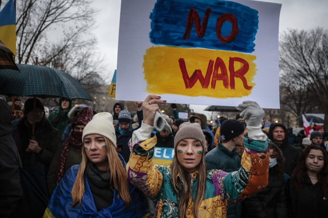 A woman in a beanie, multi-colored jacket, and face paint stands amidst a crowd of protestors holding a blue, yellow, and red sign that reads "NO WAR" 