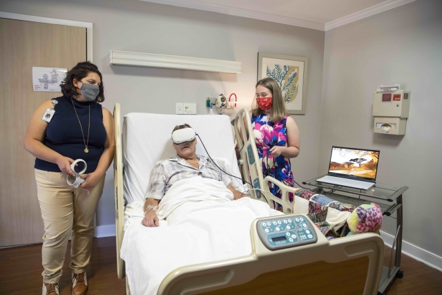 Two young women in masks and business casual work with a hospice patient wearing VR goggles, all in a warmly lit hospice room.