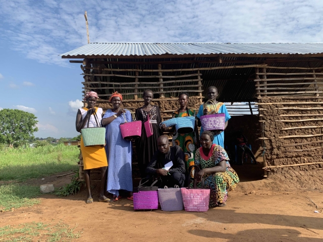 A group of women pose in a group photo holding multi-colored bags they have woven, and stand in front of a wooden cottage.