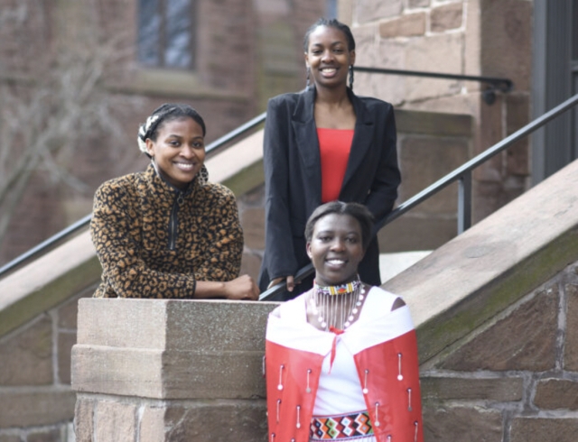Three dark-skinned young women pose for a photo on and in front of a brick stairwell.