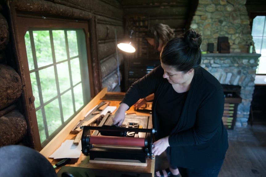 Woman stands over printing press machine within a wooden cabin