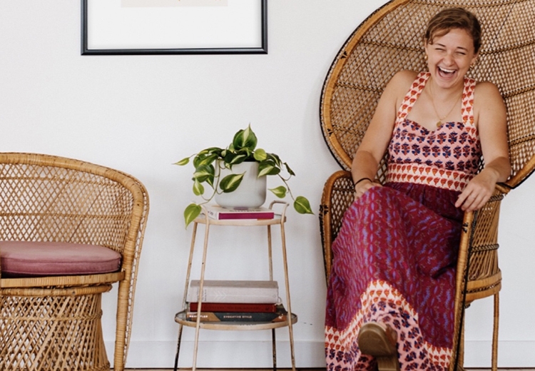 Portrait of Maddie Hoar sitting in a wicker chair next to a pothos plant