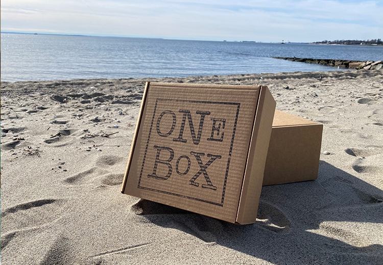 Photo of a cardboard box with the words "one box" inscribed on it rests on a beach