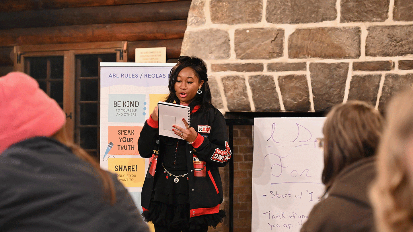 A young woman, standing, reads aloud in front of a stone fireplace.