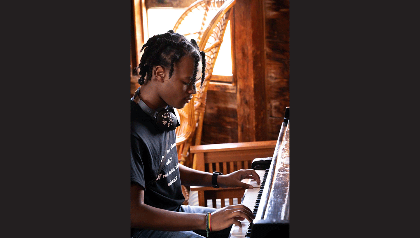 A young man plays the piano in front of a large open window. 