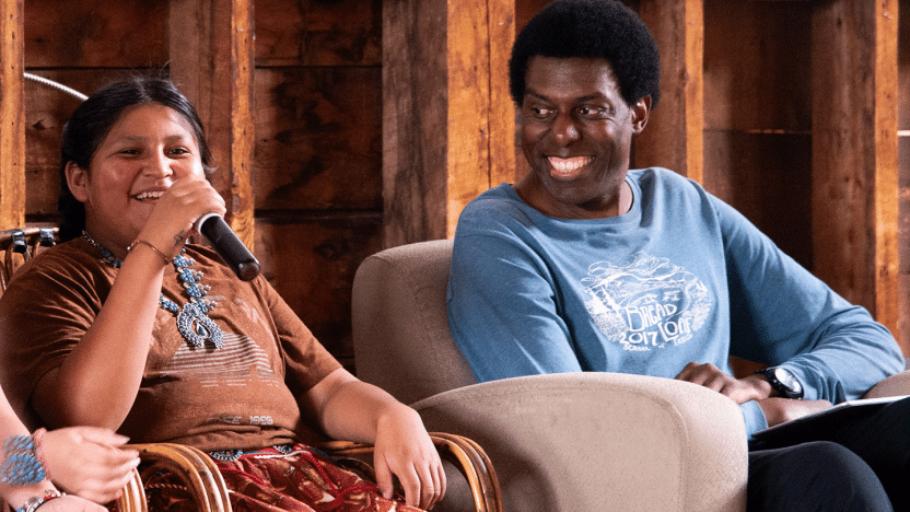 A smiling adult looks to a young woman who is speaking on a panel with a microphone. 