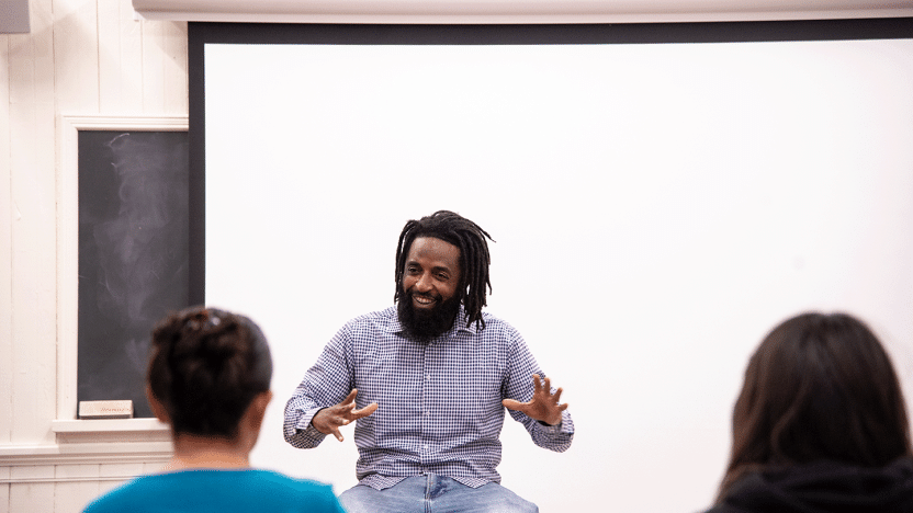 A presenter sitting on a desk smiles and gestures with both hands uplifted.