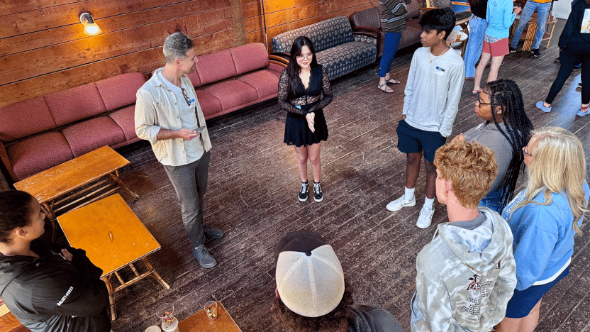 Six teens stand in a circle facing an adult actor. 