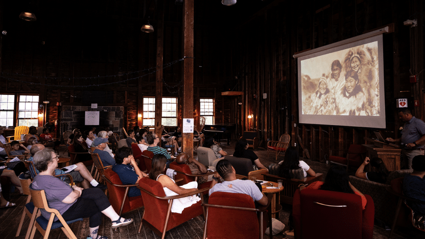 A crowd in a darkened room watches a video with an image of four Indigenous  people in fur and feather garments.