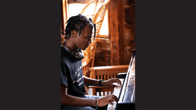 A young man plays the piano in front of a large open window. 