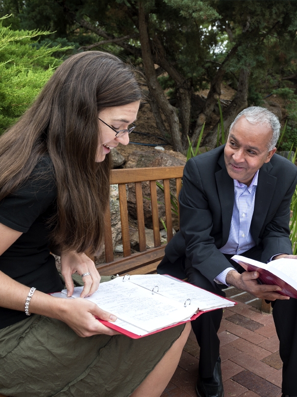 A student and professor sit outside on a wooden bench to discuss an assignment. 