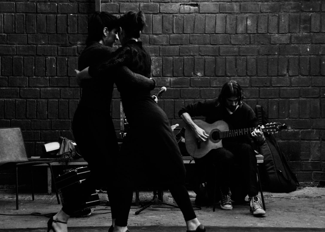 Two women dance a tango, with a guitar player seated behind them