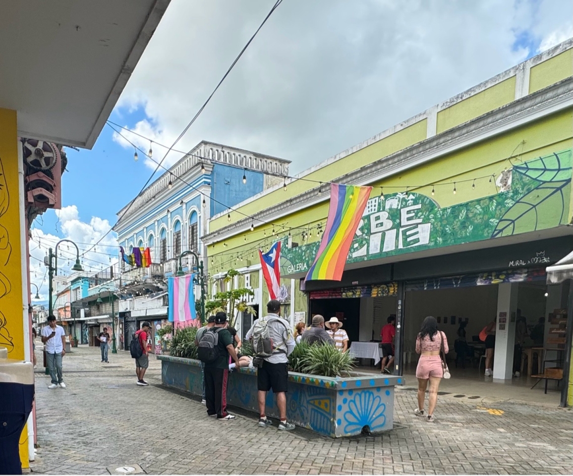 A street with people socializing. with Puerto Rican and pride flags suspended above them