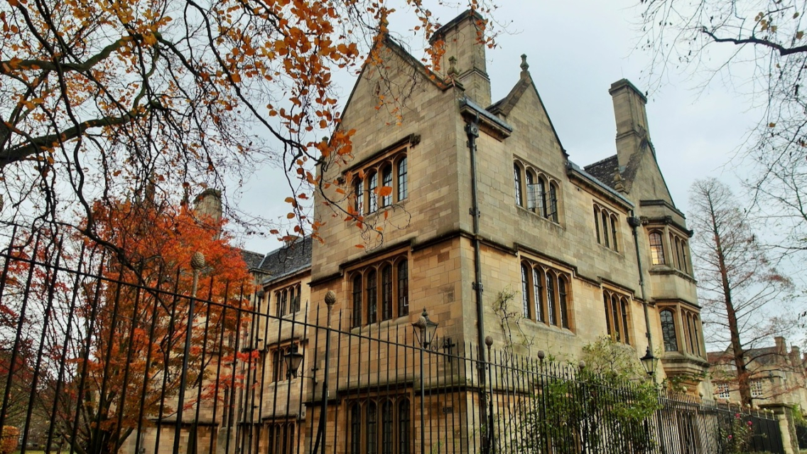 A tree with orange autumn leaves sits to the left of an Oxford college.
