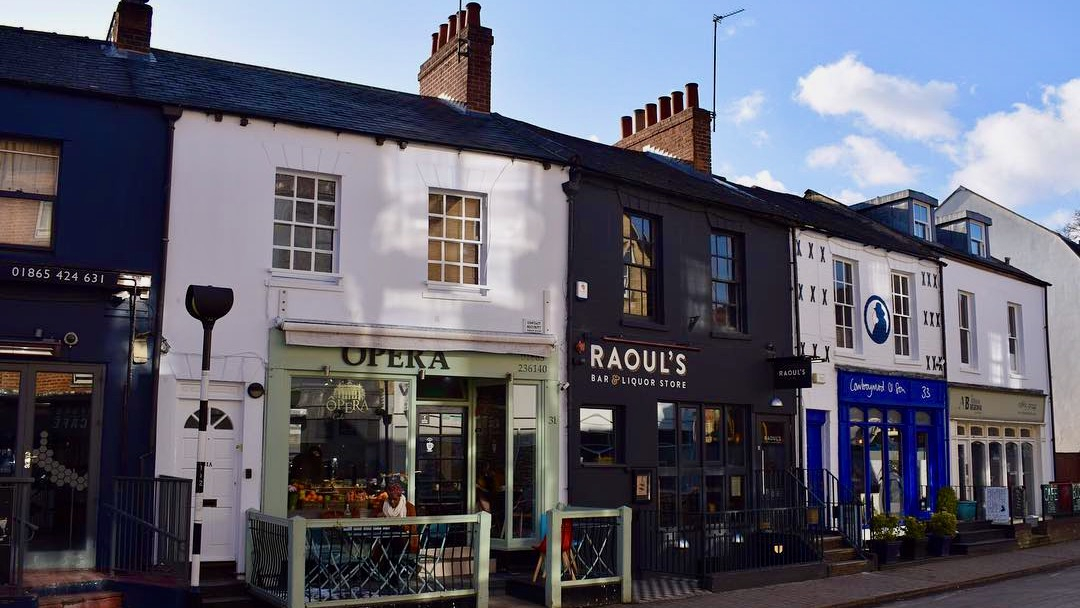 A terraced street of shops appears on a sunny day, with the Opera cafe in the foreground - a cute independent cafe that serves delicious treats.