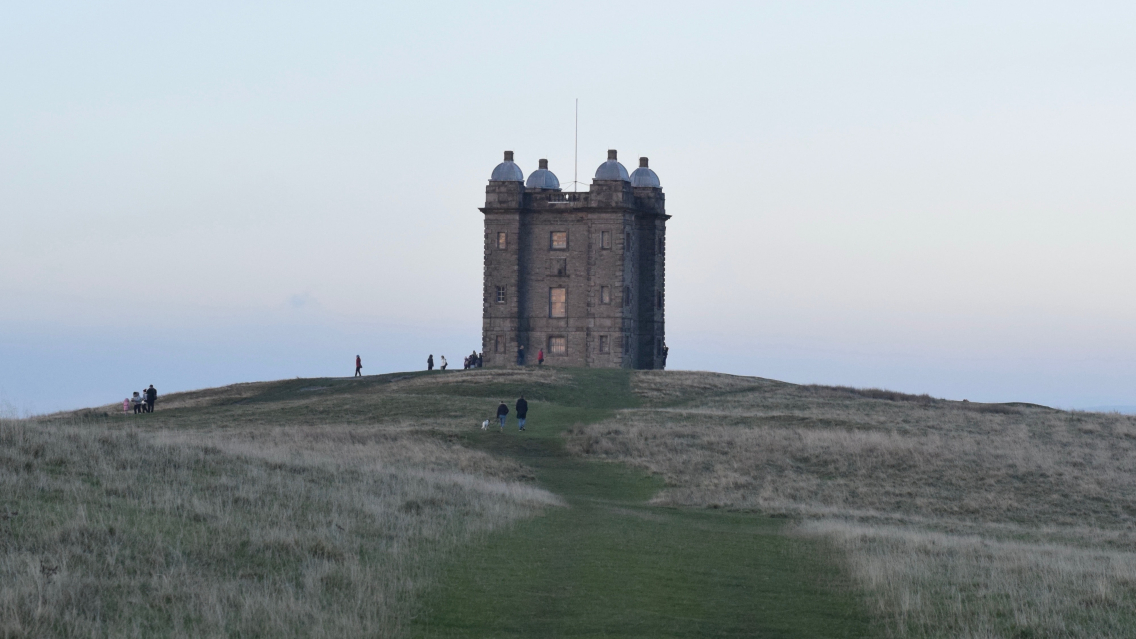 A few people are wandering along a grassy trail towards the folly at Lyme Park. It is evening, the sun is setting and the scene is bathed in a soft pastel light.