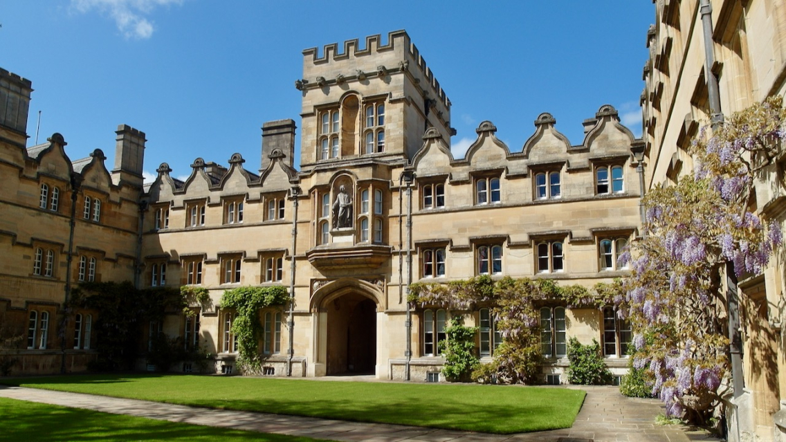 A golden stone Oxford college under a blue sky. To the right, a wisteria is in bloom with purple flowers.