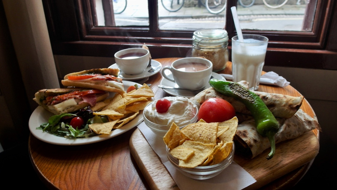 Two cups of tea and two plates piled high with food are on a table by a window in a cafe.